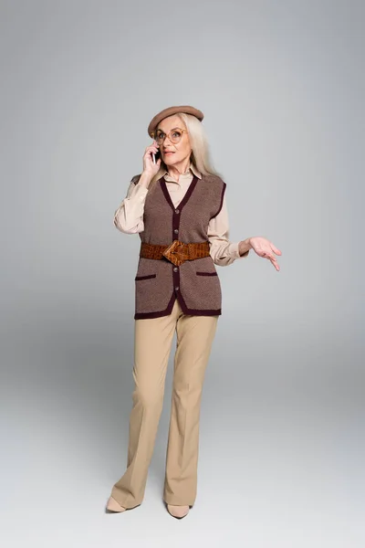 Elderly woman in beret talking on cellphone on grey background — Stock Photo