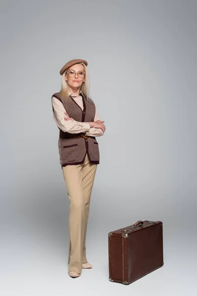 Senior woman in beret standing with crossed arms near vintage suitcase on grey background — Stock Photo