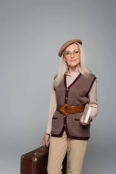 Femme sénior élégant tenant livre et valise isolé sur gris — Photo de stock