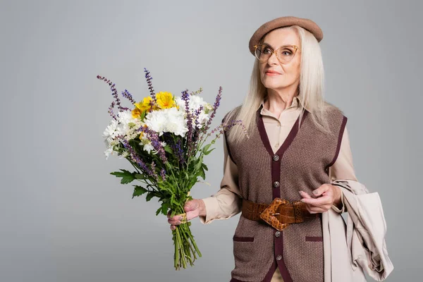 Stylish senior woman holding trench coat and bouquet isolated on grey — Stock Photo