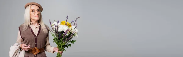 Femme âgée en béret tenant fleurs et trench coat isolés sur gris, bannière — Photo de stock