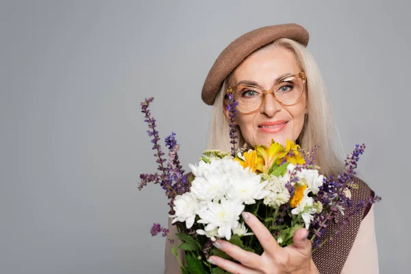 Lächelnde Seniorin in Baskenmütze und Brille mit Blumenstrauß auf Grau — Stockfoto