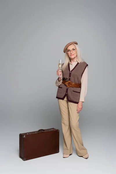 Femme âgée tenant un verre de champagne près d'une valise vintage sur fond gris — Photo de stock