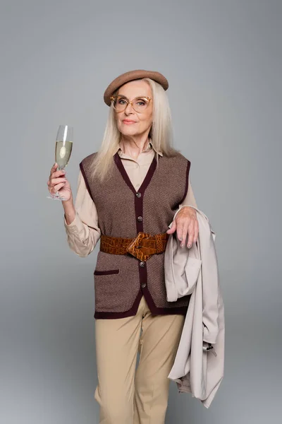 Stylish senior woman holding trench coat and glass of champagne isolated on grey — Stock Photo