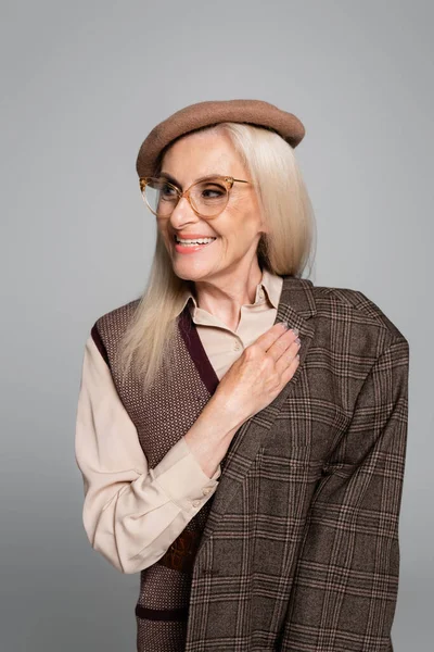 Femme âgée positive et élégante tenant veste et regardant loin isolé sur gris — Photo de stock