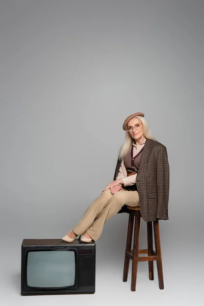 Mulher elegante sentada na cadeira perto de retro tv no fundo cinza — Fotografia de Stock