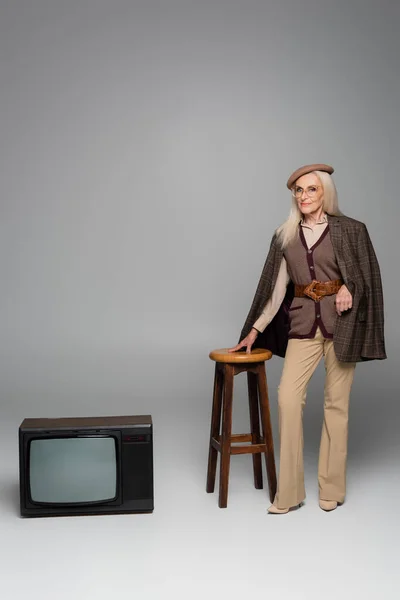 Elderly woman in beret and jacket standing near chair and vintage tv on grey background — Stock Photo