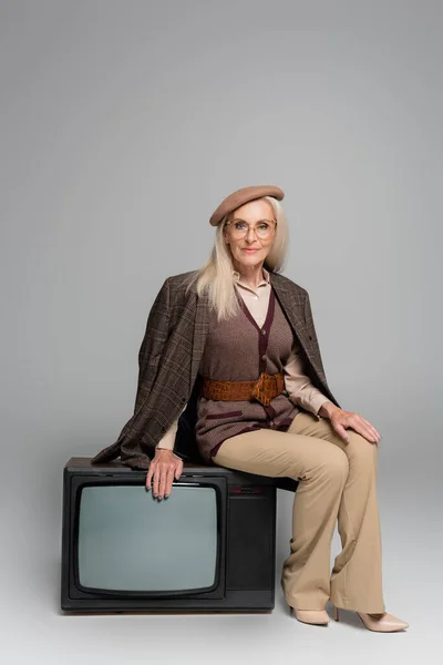 Старша жінка в плетеній куртці сидить на ретро телевізорі на сірому фоні — стокове фото