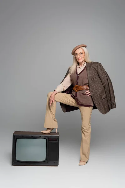 Mulher idosa de saltos altos e jaqueta de pé perto retro tv no fundo cinza — Fotografia de Stock