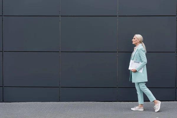 Vista laterale della donna d'affari anziana con giornale che cammina vicino all'edificio all'aperto — Foto stock