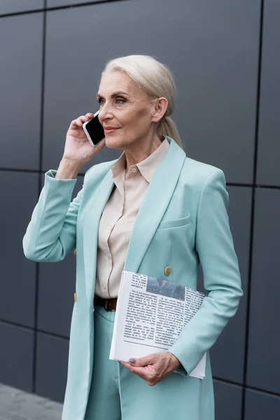 Empresa sénior sosteniendo periódico y hablando en un teléfono inteligente cerca del edificio al aire libre - foto de stock