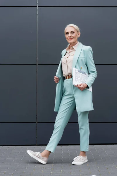 Femme d'affaires senior avec journal regardant la caméra près du bâtiment à l'extérieur — Photo de stock