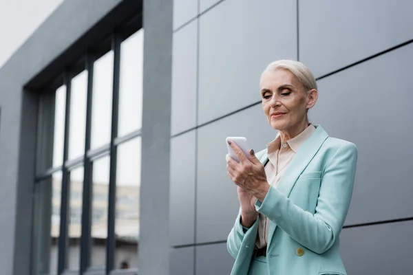 Elderly businesswoman using cellphone while standing near building outdoors — Stock Photo