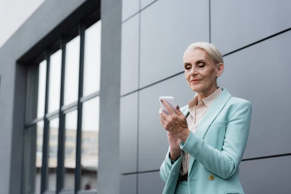 Mujer de negocios de edad avanzada usando el teléfono celular mientras está de pie cerca del edificio al aire libre — Stock Photo