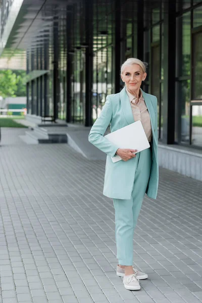 Seniorin hält Laptop in der Hand und blickt auf Kamera in der Nähe von Haus im Freien — Stockfoto