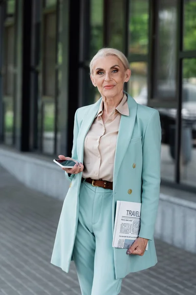 Femme d'affaires senior tenant smartphone et journal en plein air — Photo de stock