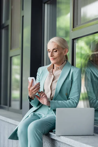Elderly businesswoman using smartphone near laptop and building outdoors — Stock Photo