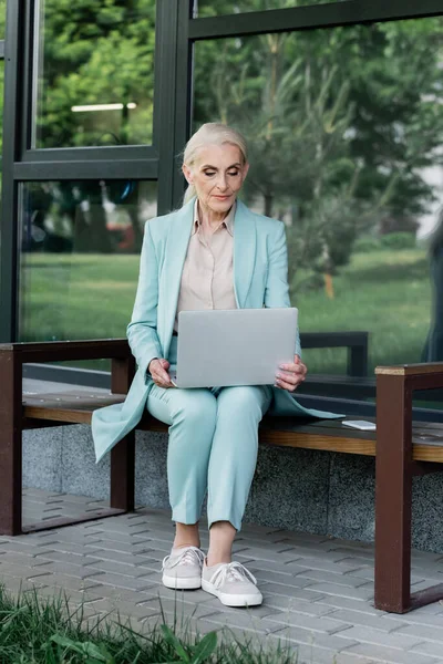 Anciana mujer de negocios utilizando el ordenador portátil cerca de teléfono inteligente en el banco - foto de stock