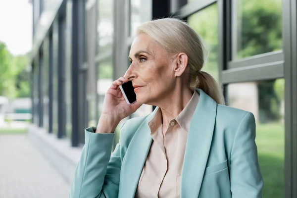 Vista lateral de la mujer de negocios senior hablando por teléfono móvil cerca del edificio borroso - foto de stock