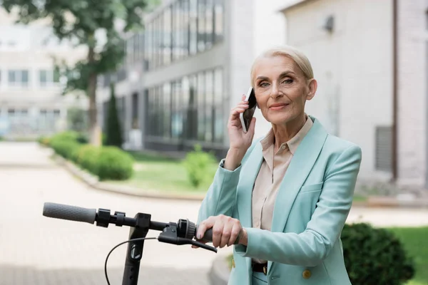 Elderly businesswoman talking on mobile phone near electric kick scooter outdoors — Stock Photo