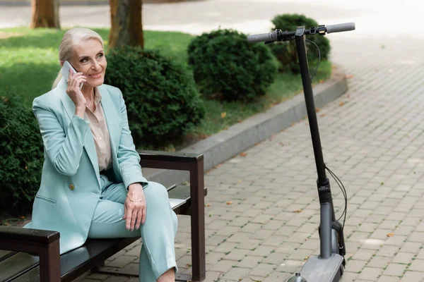 Joyful businesswoman calling on mobile phone on bench near electric kick scooter — Stock Photo