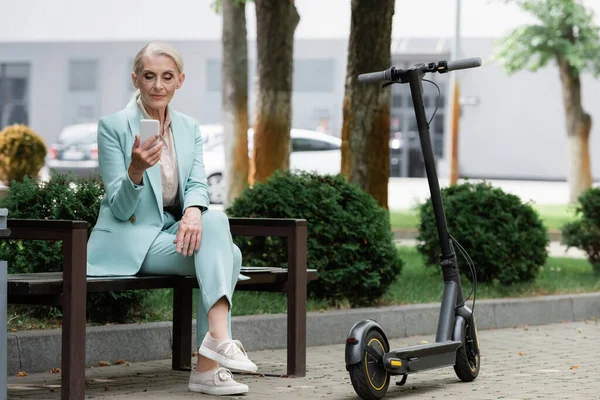 Seniorin benutzt Smartphone, während sie auf Bank neben Elektro-Tretroller sitzt — Stockfoto