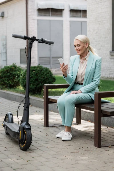 Senhora de negócios sênior elegante sentado no banco com smartphone perto de scooter de chute elétrico — Fotografia de Stock