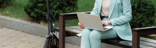 Visão parcial da mulher idosa usando laptop no banco perto de scooter de chute elétrico, banner — Fotografia de Stock