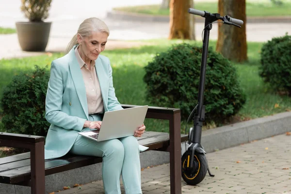 Ältere Frau benutzt Laptop auf Bank in der Nähe von Elektro-Tretroller — Stockfoto
