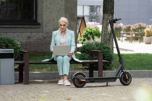 Seniorin im blauen Anzug benutzt Smartphone, während sie auf Bank neben Roller sitzt — Stockfoto
