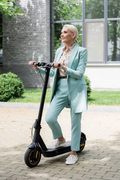 Mujer de negocios senior de pie en la calle con patinete scooter eléctrico y mirando hacia otro lado - foto de stock