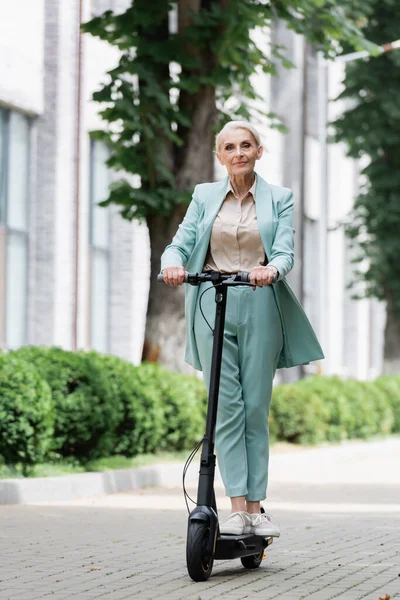 Anciana mujer de negocios en traje azul montando patinete eléctrico scooter al aire libre - foto de stock