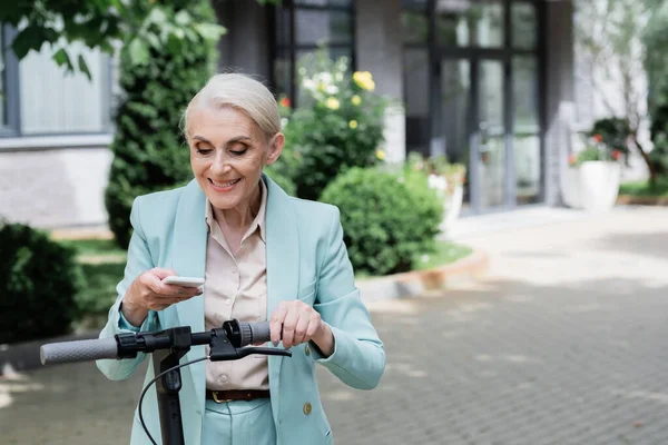 Senior-Geschäftsfrau lächelt beim Benachrichtigen auf Smartphone in der Nähe von Elektro-Tretroller — Stockfoto