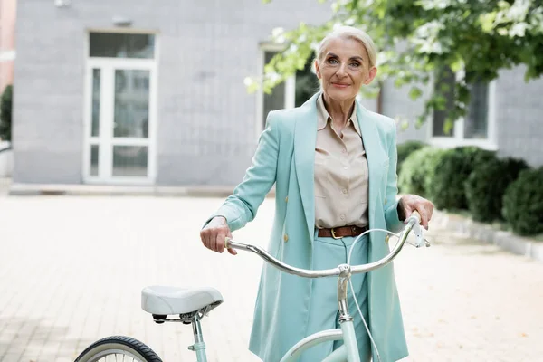 Stylish senior businesswoman smiling at camera near bicycle outdoors — Stock Photo