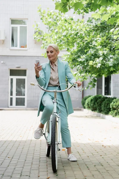 Старшая бизнес-леди с помощью мобильного телефона, сидя на велосипеде на открытом воздухе — стоковое фото