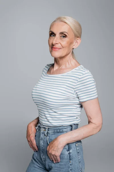 Grey haired woman in striped t-shirt posing with thumbs in pockets of jeans isolated on grey — Stock Photo