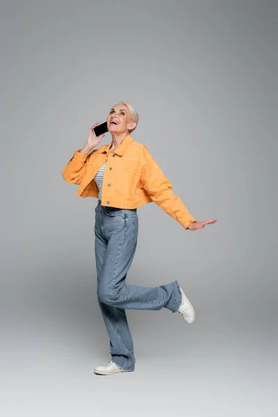 Mujer mayor emocionada y elegante hablando en el teléfono inteligente mientras está de pie en una pierna en gris - foto de stock