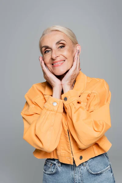 Cheerful senior woman in orange jacket posing with hands near face isolated on grey — Stock Photo