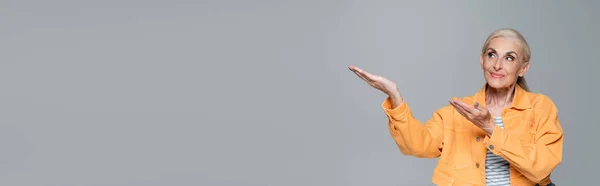 Positive elderly woman in orange jacket looking away and pointing with hands isolated on grey, banner — Stock Photo