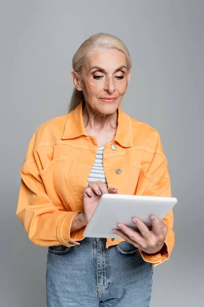 Старшая женщина в оранжевой куртке с помощью цифровой таблетки изолированы на серый — стоковое фото