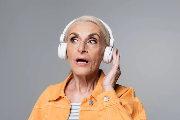 Astonished elderly woman adjusting headphones while looking away isolated on grey — Stock Photo