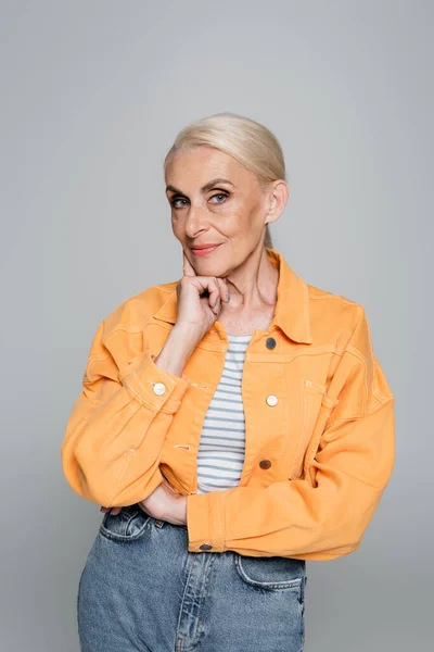 Elderly woman in trendy jacket holding hand near face isolated on grey — Stock Photo