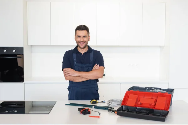 Reparador sonriente de pie con los brazos cruzados cerca de herramientas y cables en la cocina - foto de stock