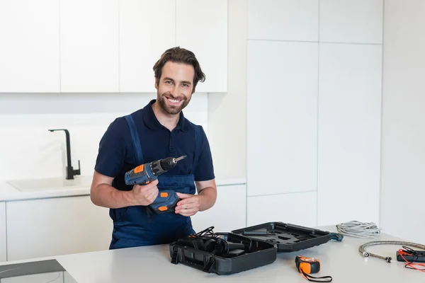 Glücklicher Arbeiter mit Elektrobohrmaschine blickt auf Kamera in der Nähe von Werkzeugen auf Küchentisch — Stockfoto