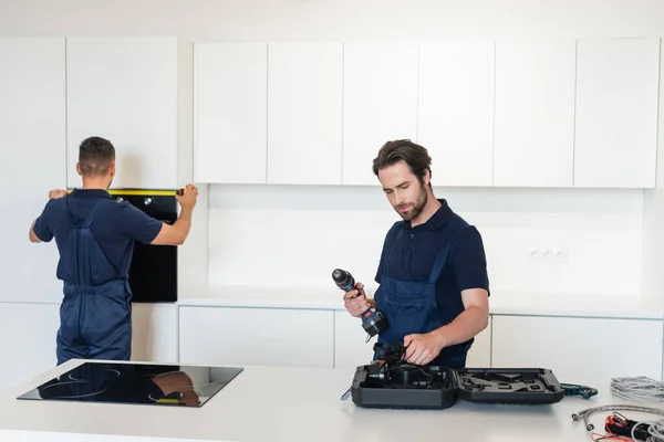 Arbeiter hält Elektrobohrer neben Kollegen und misst eingebauten Ofen in Küche — Stockfoto