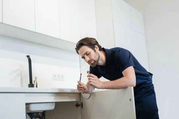 Klempner in Uniform hält Metallschlauch nahe Spüle in Küche — Stockfoto