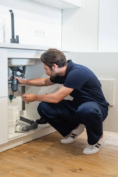 Plumber in uniform fixing metal hose under kitchen sink — Stock Photo