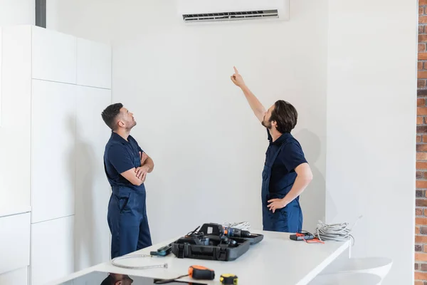 Reparador apuntando al aire acondicionado cerca de colega y herramienta en la mesa de la cocina - foto de stock