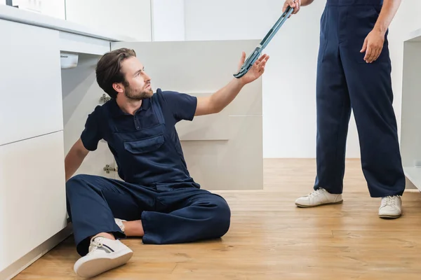 Arbeiter gibt Klempner, der in Küche auf dem Boden sitzt, einen verstellbaren Schraubenschlüssel — Stockfoto