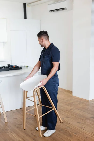 Foreman looking at electric drill while holding white stool in kitchen — Stock Photo